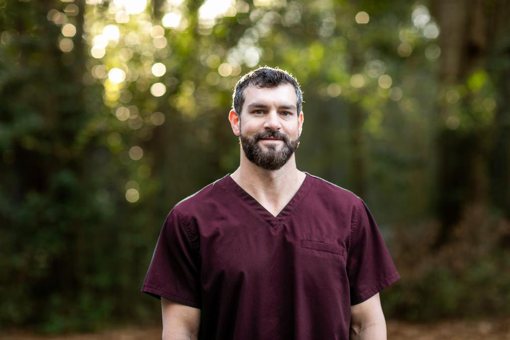 A doctor with dark hair and a beard in maroon scrubs standing outside in a natural green environment